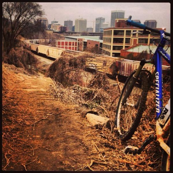 A rugged bike rests on a dirt path overlooking a train track, with the skyline of a city in the background, featuring tall buildings and a cloudy sky. The scene captures a blend of nature and urban landscape. Northbank Trail mountain bike trail.