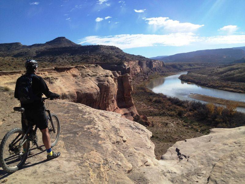 A mountain biker standing on a rocky ledge overlooking a river valley, with mountains in the background under a partly cloudy blue sky. The scene captures a sense of adventure and the beauty of nature. Kokopelli Area Trails mountain bike trail.
