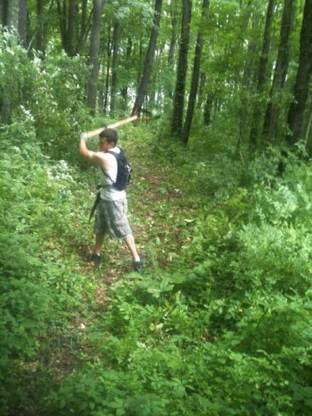 A person standing on a trail in a lush green forest, swinging a stick overhead. The individual is wearing a tank top and cargo shorts, with a backpack on their back. Surrounding vegetation includes trees and dense undergrowth, creating a vibrant outdoor scene. Barkcamp State Park mountain bike trail.