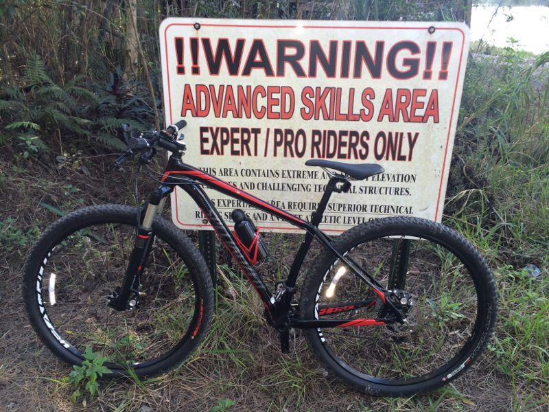 A mountain bike is leaning against a large warning sign that reads, "WARNING!! ADVANCED SKILLS AREA EXPERT/PRO RIDERS ONLY." The sign details that the area contains extreme elevation changes and challenging technical structures, indicating that superior technical skills and athletic ability are required for riding in this area. Surrounding the sign is dense vegetation. Markham Park mountain bike trail.