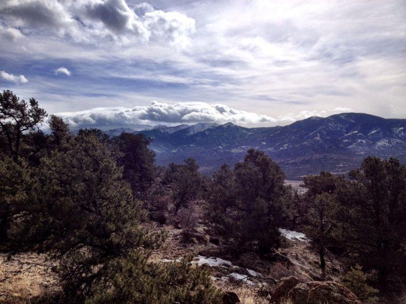 A panoramic view of mountains under a partly cloudy sky, surrounded by evergreen trees in the foreground. Snow is visible on the ground, indicating a cold climate. The landscape showcases a mix of rugged terrain and cloud formations, creating a serene and picturesque natural scene. Pauli mountain bike trail.