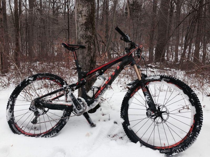 A mountain bike with a black and red frame is leaning against a tree in a snowy forest. The bike is partially covered in snow, showcasing its tires and suspension. Surrounding trees are bare, indicating winter conditions. Hartshorne Woods Park mountain bike trail.