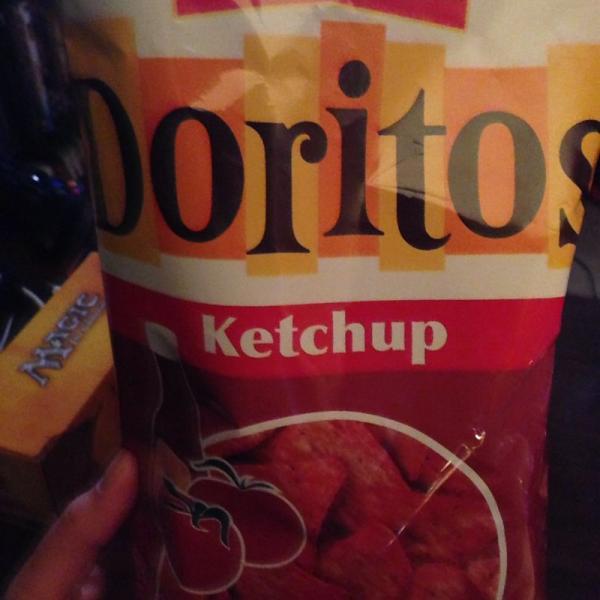 A close-up image of a bag of Doritos chips featuring a ketchup flavor. The bag has a yellow and red color scheme with the word "Ketchup" prominently displayed beneath the Doritos logo. A hand is partially visible, holding the bag. The Pines mountain bike trail.