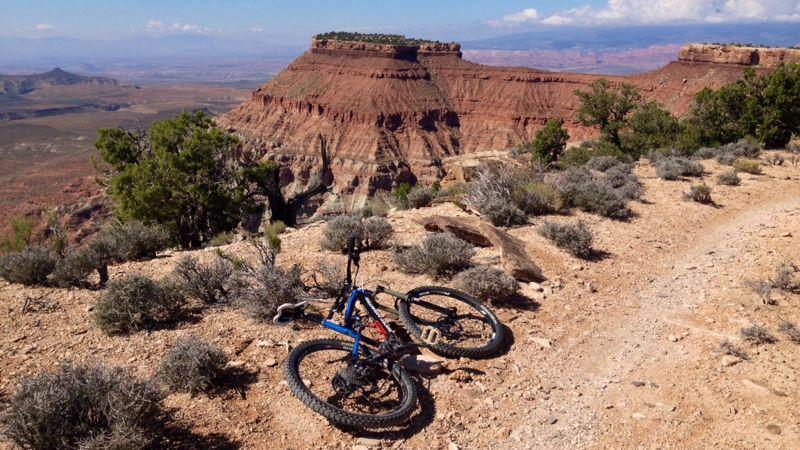A mountain bike rests on rocky terrain, overlooking a vast canyon landscape. In the background, distinctive red rock formations and distant mountains are visible under a clear blue sky with a few clouds. Sparse vegetation surrounds the bike, highlighting the rugged outdoor environment. Gooseberry Mesa mountain bike trail.