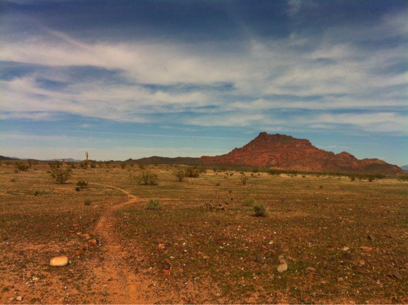 A vast, arid landscape featuring a dusty trail winding through sparse vegetation, with a prominent reddish-brown mountain in the background under a partly cloudy sky. Hawes Loop mountain bike trail.