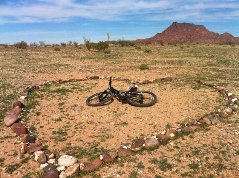 A black bicycle lying on the ground inside a heart-shaped arrangement of stones, situated in a rocky desert landscape with a distant mountain and blue sky. Hawes Loop mountain bike trail.
