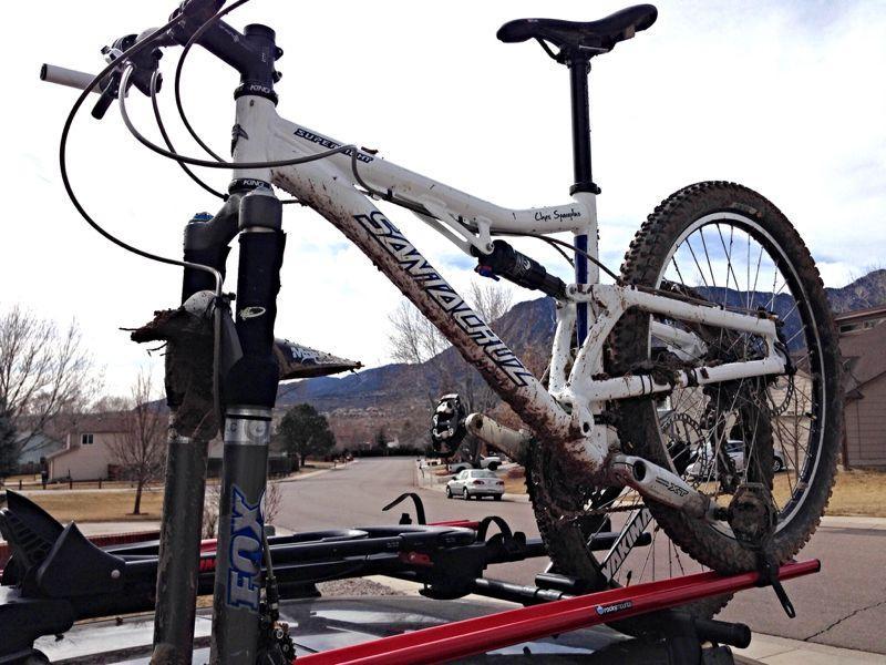 A white mountain bike mounted on the roof of a vehicle, showing muddy tires and components. The background features a residential area with trees and mountains in the distance. Cheyenne Mountain State Park mountain bike trail.