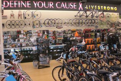 A bicycle store interior featuring various bike frames, accessories, and equipment displayed on shelves and racks. The back wall prominently displays the phrases "DEFINE YOUR CAUSE" and "LIVESTRONG" along with circular bike parts. The shop has a bright and organized layout, showcasing a variety of bicycles in the foreground.