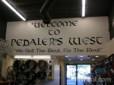 Sign featuring the text "Welcome to Pedaler's West" with the slogan "We Sell The Best, Fix The Rest" underneath. The sign is displayed prominently in a shop interior, with bicycle accessories and tires visible in the background.