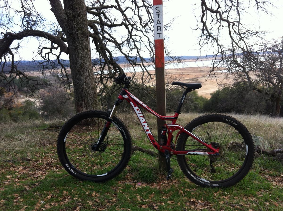A red mountain bike is leaning against a wooden post marked "START," situated in a grassy area surrounded by trees. In the background, a scenic view of a body of water and rolling hills is visible, with a cloudy sky overhead. Granite Bay Trail mountain bike trail.