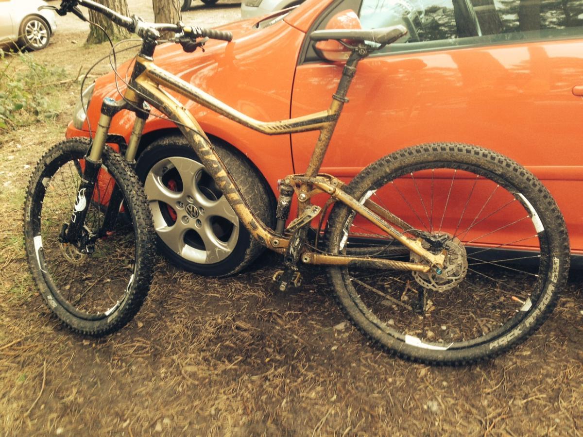 Giant Trance SX 27.5: A mountain bike with a muddy frame and tires is leaning against an orange car. The bike has a sleek design, featuring wide tires suitable for rough terrain and a suspension system. The background shows a natural setting with trees and gravel.