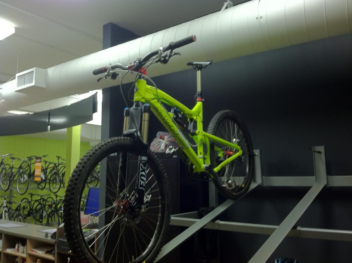 Santa Cruz Nomad: A bright green mountain bike is displayed on a rack in a bike shop, showcasing its front wheel and suspension. The background features a wall of bicycles and a counter with various items. The setting appears well-lit and organized, emphasizing the bike's vibrant color and design.