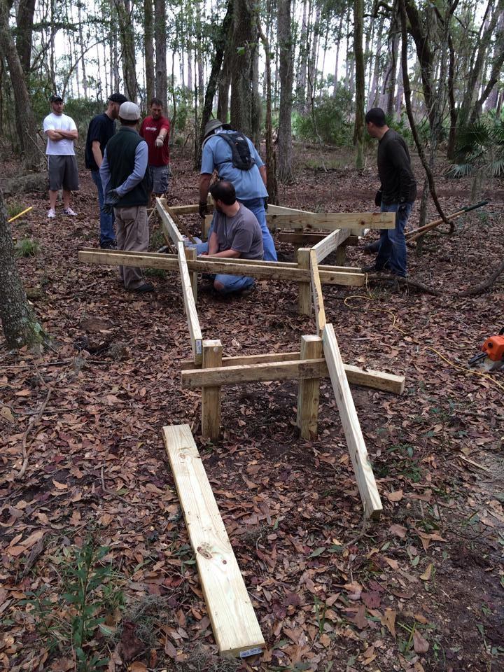 A group of people working together in a wooded area, assembling a wooden structure. They are surrounded by fallen leaves, with several wooden beams and tools scattered around. Some individuals are discussing or pointing, while others are focused on the construction process. Tall trees and greenery are visible in the background. Nocatee mountain bike trail.