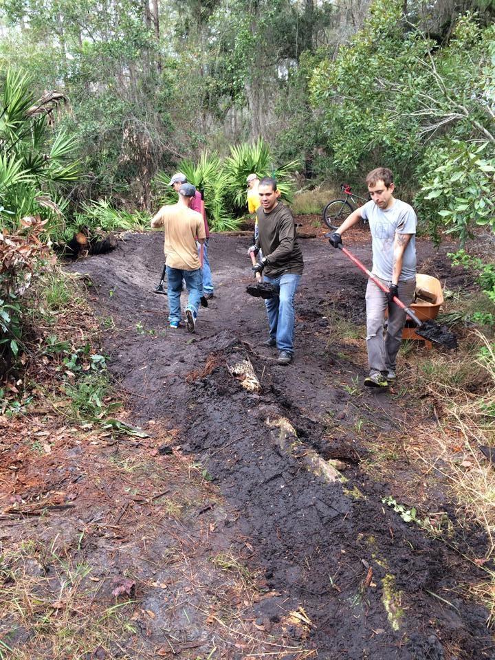 A group of five individuals working on a muddy trail in a natural setting, equipped with tools like shovels and a rake. They are engaged in outdoor maintenance, surrounded by greenery and trees, with some participants wearing casual clothing. One person is pushing a wheelbarrow. Nocatee mountain bike trail.