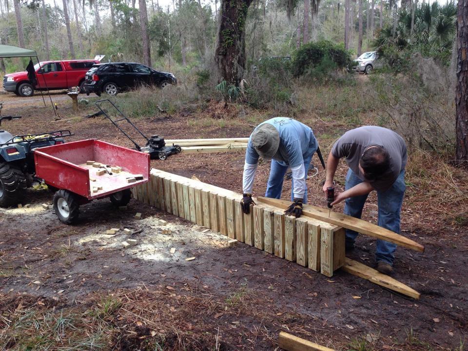 Two men working together to construct a wooden structure in a forested area. One man is holding a piece of wood while the other is using a tool. Nearby, there's an ATV with a red cart filled with wooden pieces, and additional lumber is visible on the ground. Trees and vehicles are visible in the background, indicating an outdoor project setting. Nocatee mountain bike trail.