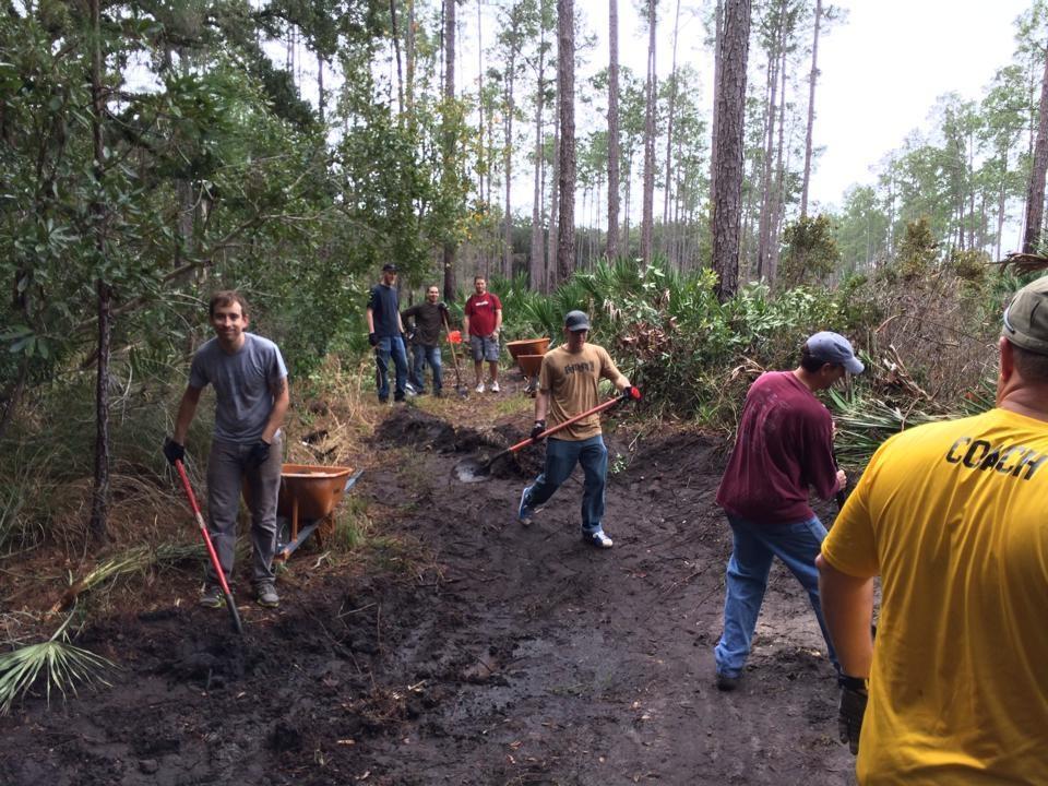 A group of people working together to clear a muddy trail in a wooded area. They are using shovels and wheelbarrows, with trees and plants visible in the background. The scene captures collaborative outdoor activity focused on environmental maintenance. Nocatee mountain bike trail.