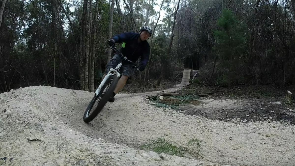 A mountain biker navigating a dirt trail with a banked turn in a forest setting. The biker is wearing a helmet and casual riding gear, showcasing a moment of dynamic motion on a mountain bike. The surrounding environment features trees and rough terrain, indicating an outdoor biking trail. Tillie Fowler Regional Park mountain bike trail.