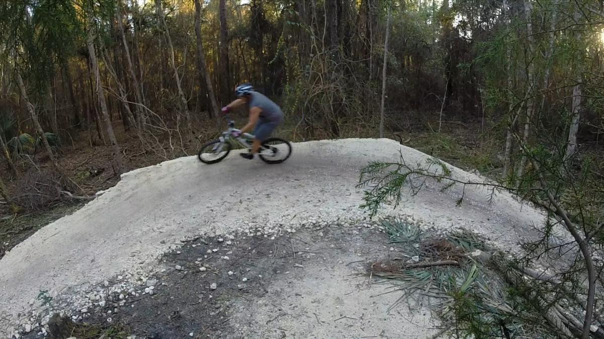 A cyclist riding a mountain bike along a curved dirt path in a wooded area, with trees and underbrush in the background. Tillie Fowler Regional Park mountain bike trail.