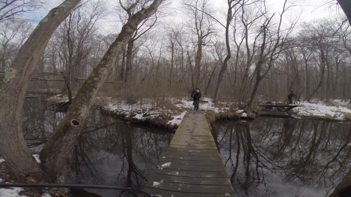 A narrow wooden bridge spanning a dark, reflective stream in a wooded area during winter. Leafless trees surround the scene, and patches of snow are visible on the ground. A person is walking on the bridge while another person is riding a bike nearby. The sky is overcast, creating a muted lighting effect. Mercer County Park mountain bike trail.