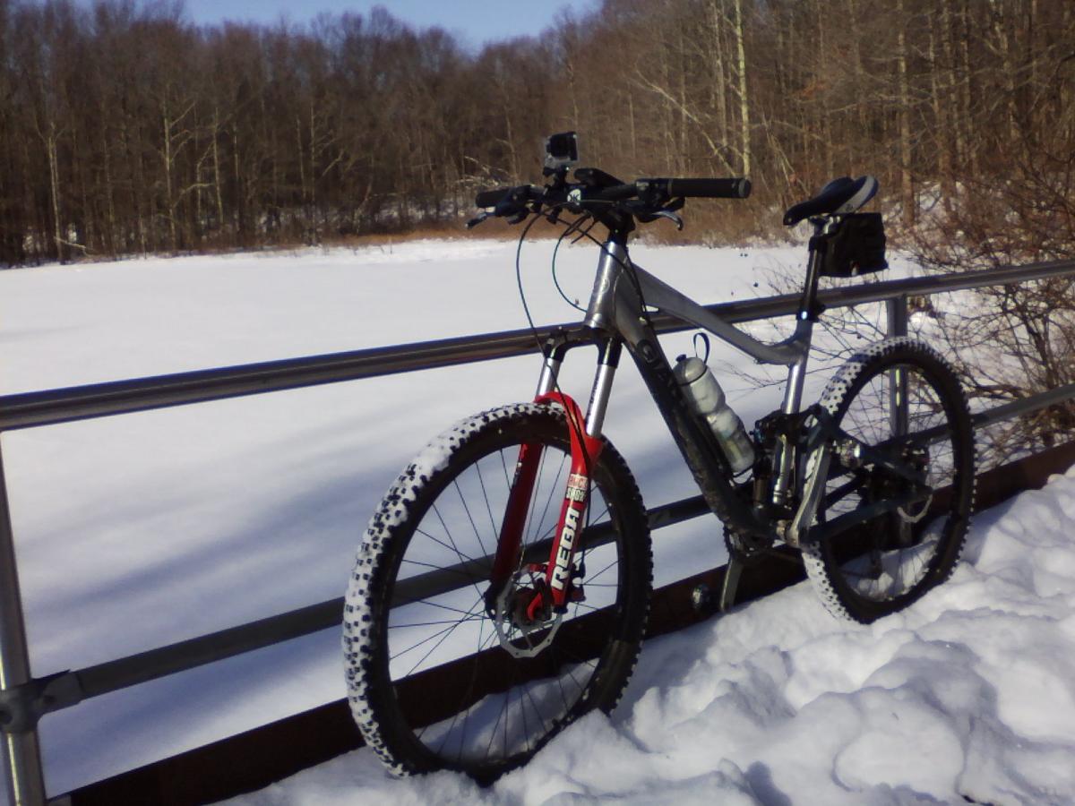 A mountain bike with a black and red frame is parked on a snow-covered path beside a frozen lake, surrounded by bare trees. The scene captures a tranquil winter landscape with clear blue skies. Wolfes Pond park mountain bike trail.