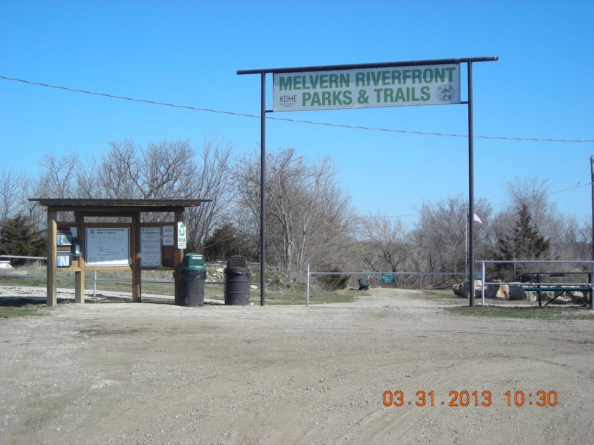 Entrance to Melvern Riverfront Parks & Trails featuring a sign overhead, information board, and waste bins. The background includes bare trees against a clear blue sky. The area is surrounded by gravel and provides access to recreational activities. Melvern Riverfront Trails mountain bike trail.