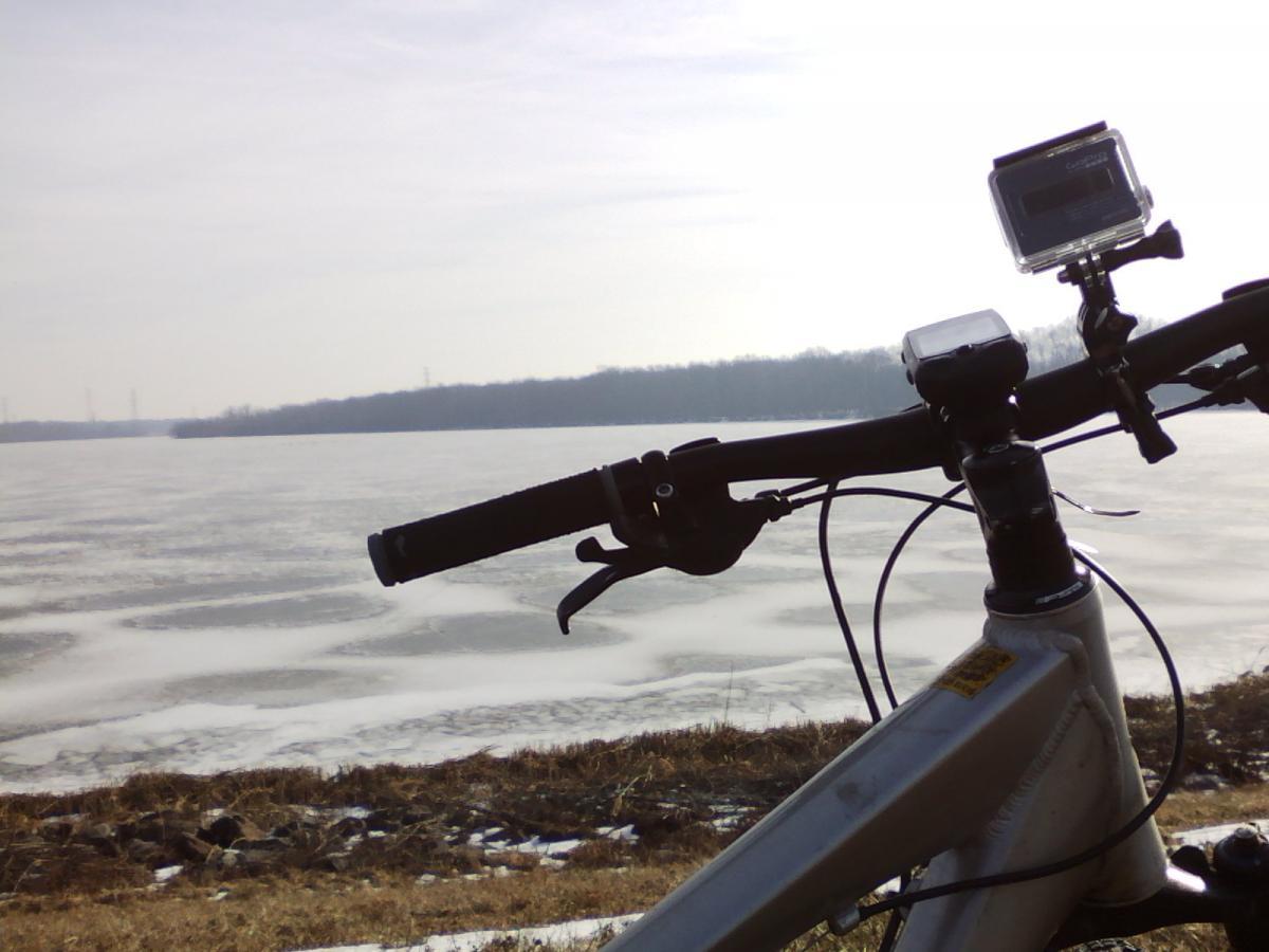 A close-up view of a bicycle's handlebars positioned next to a frozen body of water. In the background, there are bare trees and a cloudy sky, suggesting a chilly environment. A camera is mounted on the handlebars, indicating a potential for recording or filming the scenic view. The ground is partially covered with grass and remnants of snow. Mercer County Park mountain bike trail.