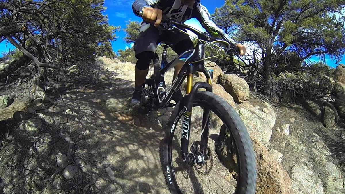 A mountain biker navigating a rocky trail under a blue sky with scattered clouds, surrounded by trees. The cyclist is wearing protective gear and is balancing on the bike as it climbs over uneven terrain. Unkle Nazty mountain bike trail.