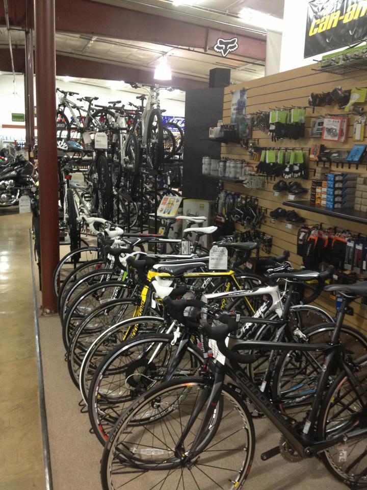 A bike shop interior featuring a row of various bicycles displayed prominently in the foreground. In the background, additional bicycles are visible on racks, along with shelves stocked with biking accessories, tools, and apparel. The store has high ceilings with overhead lighting and a clean, organized layout.