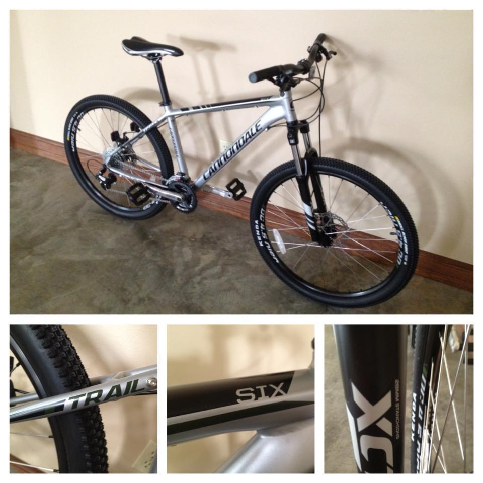 A silver Cannondale mountain bike is leaning against a wall. The bike features a sturdy frame, thick tires, and disc brakes, with close-up views of the "Trail" and "STX" labels on the frame. The setting has a concrete floor and a neutral-colored wall.