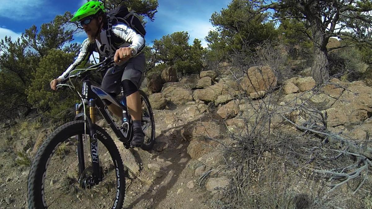 A mountain biker riding on a rocky trail surrounded by trees, wearing a green helmet and athletic gear. The bike is navigating a challenging path with large stones and sparse vegetation under a blue sky. Unkle Nazty mountain bike trail.