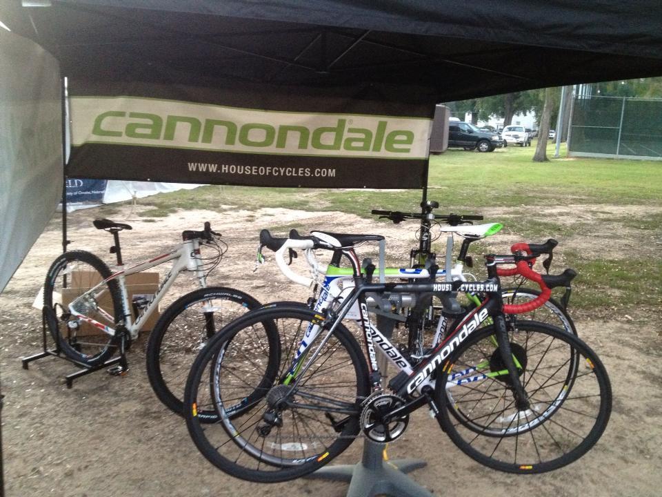 A display of Cannondale bicycles under a tent, featuring a black road bike with green accents in the foreground, alongside a white mountain bike in the background. The tent displays the Cannondale logo and the website "www.houseofcycles.com." The setting is outdoors, with grass and trees visible in the background.