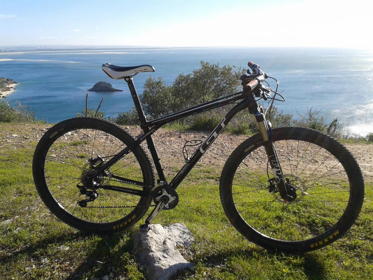 GT kashmir: A mountain bike positioned on a grassy hill overlooking a tranquil body of water, with distant hills and an island visible in the background. The scene captures a clear sky and sunlight reflecting on the water