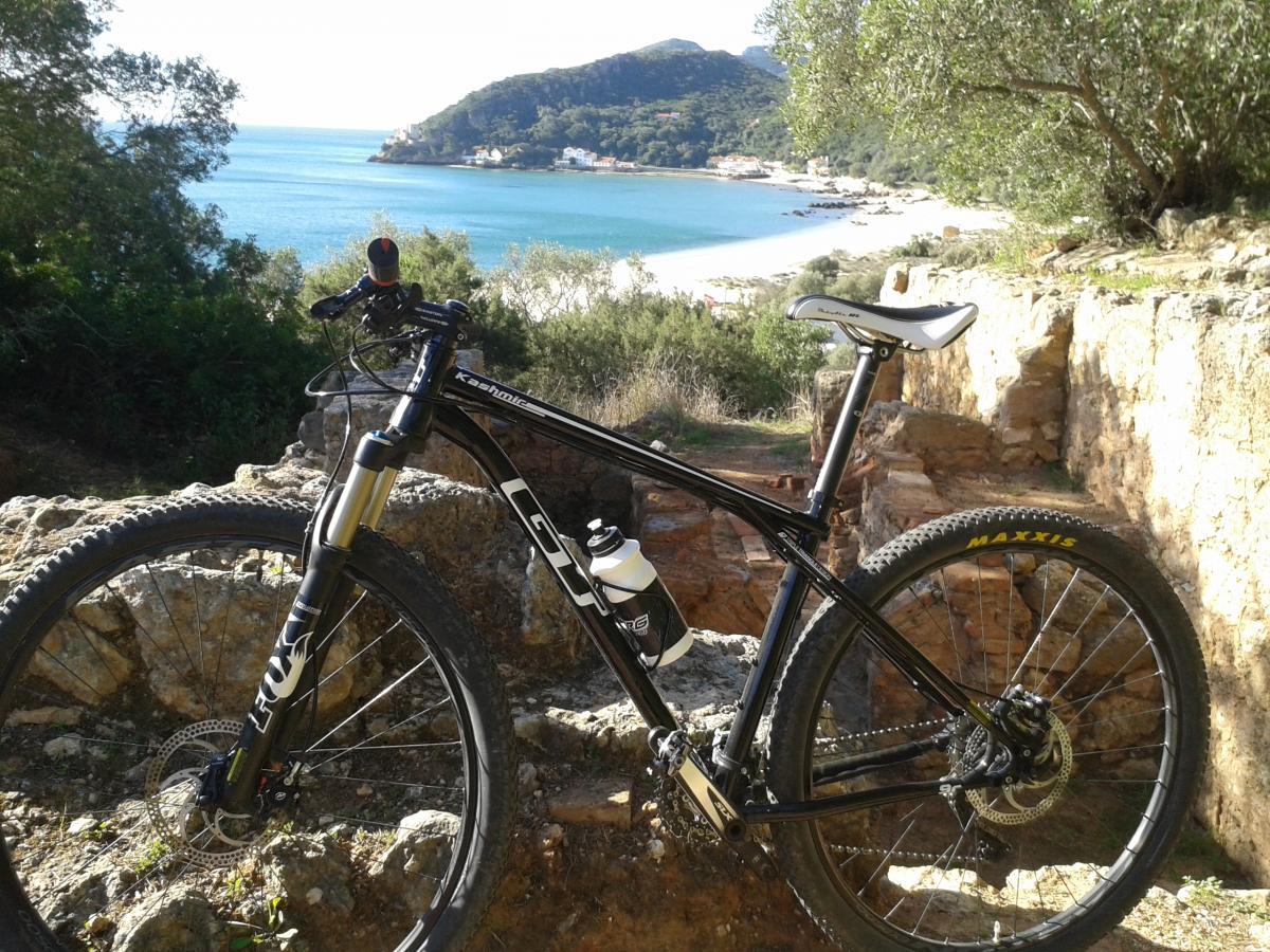 GT kashmir: A black mountain bike with a white seat and water bottle is parked on a rocky ledge, overlooking a scenic view of a blue bay and a sandy beach surrounded by lush green hills. The sunlight casts a warm glow over the landscape, highlighting the bike and the beautiful coastal setting.