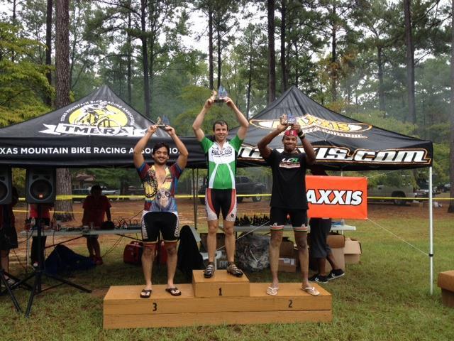 Three mountain bikers stand on a podium celebrating their race results. The first-place winner in green and white gear holds a trophy, flanked by the second-place winner in black and the third-place winner in colorful attire. Behind them is a large, branded tent set up for the event in a forested area.
