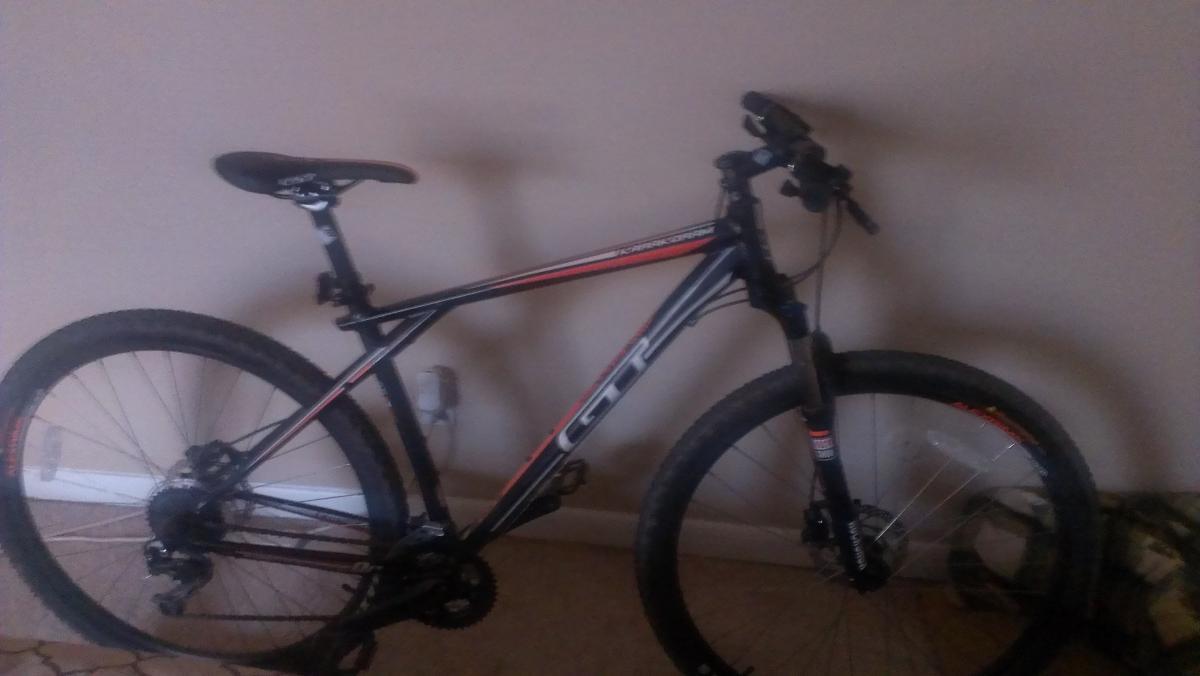 GT Karakoram: A black and red mountain bike leaning against a wall, showcasing its frame, handlebars, and wheels. The background is a plain wall with a carpeted floor.