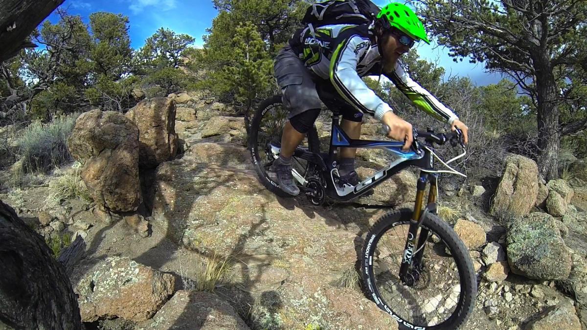 A mountain biker in a bright green helmet and athletic gear navigates rocky terrain, leaning forward on a blue and black mountain bike. Sunlight shines on the surrounding trees and rocky landscape, emphasizing the adventurous outdoor setting. Unkle Nazty mountain bike trail.