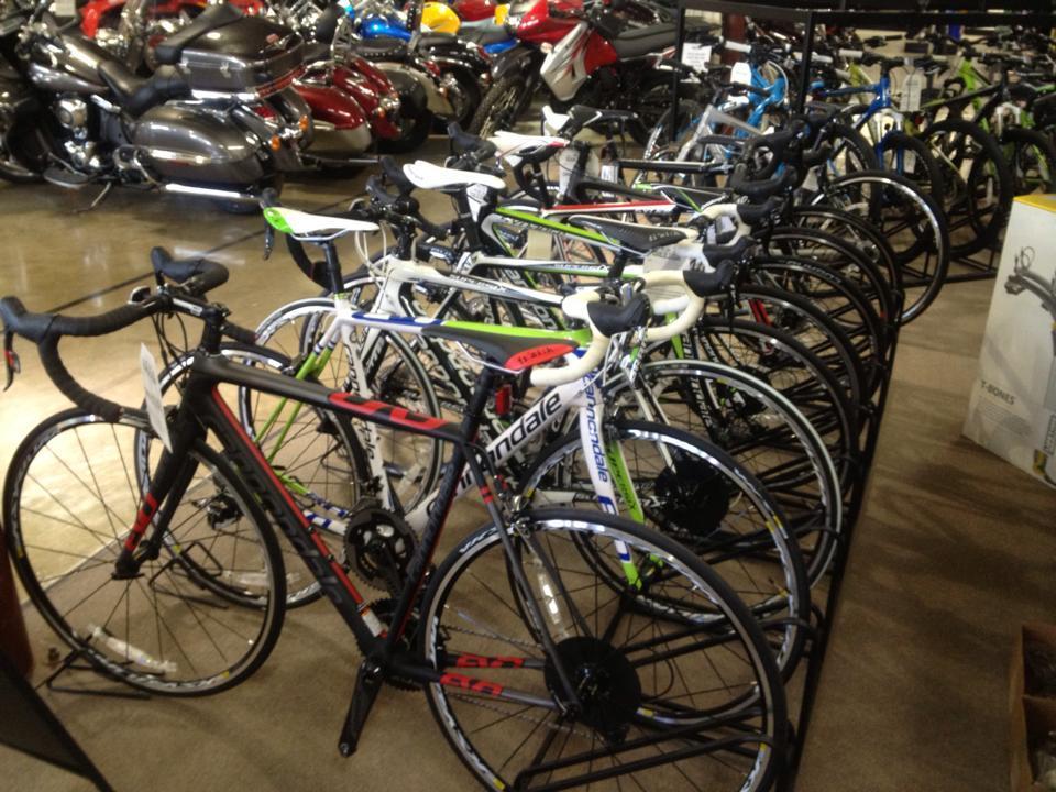 A display of various road bicycles in a bike shop, featuring different colors and designs, with several bikes lined up on a rack. In the background, multiple motorcycles can be seen, indicating a diverse selection of vehicles available in the store.