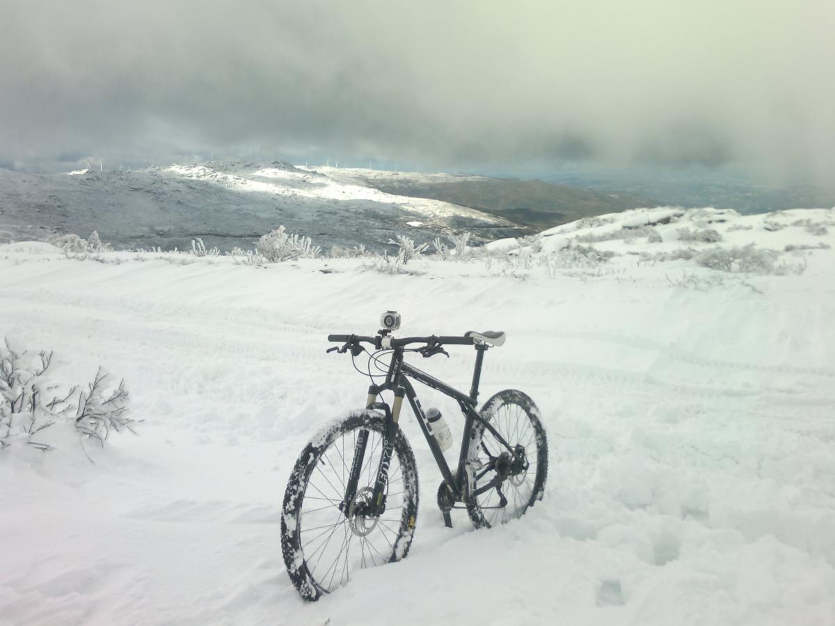 GT kashmir: A mountain bike resting on a snowy trail with a winter landscape in the background, featuring rolling hills and a cloudy sky. The bike has a camera mounted on it, and snow blankets the ground and nearby vegetation.