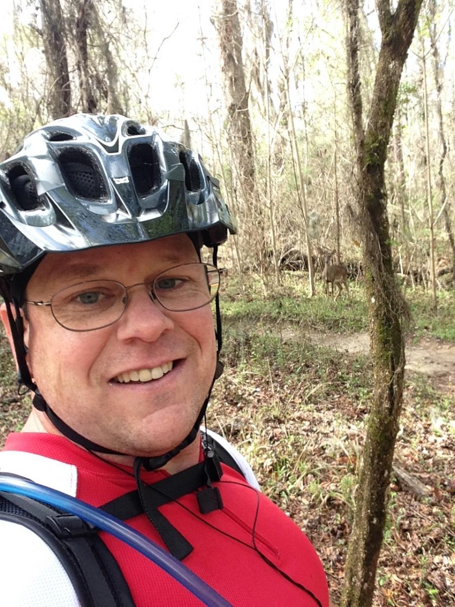 A smiling person wearing a black bicycle helmet and a red shirt stands on a wooded trail, with trees in the background and a deer visible in the distance. The person is holding a hydration pack and appears to be enjoying a cycling or hiking adventure. San Felasco Hammock Preserve mountain bike trail.