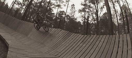 A cyclist navigating a wooden pump track in a forested area, featuring curves and elevation changes, captured in a black-and-white tone. Blankets Creek mountain bike trail.