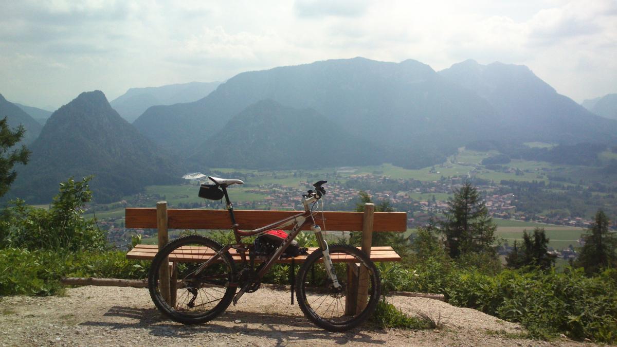 Trek Fuel EX 8: A mountain bike parked next to a wooden bench, overlooking a scenic view of mountains and a valley with a small town below. The sky is partly cloudy, and the landscape features lush greenery and rugged peaks in the background.