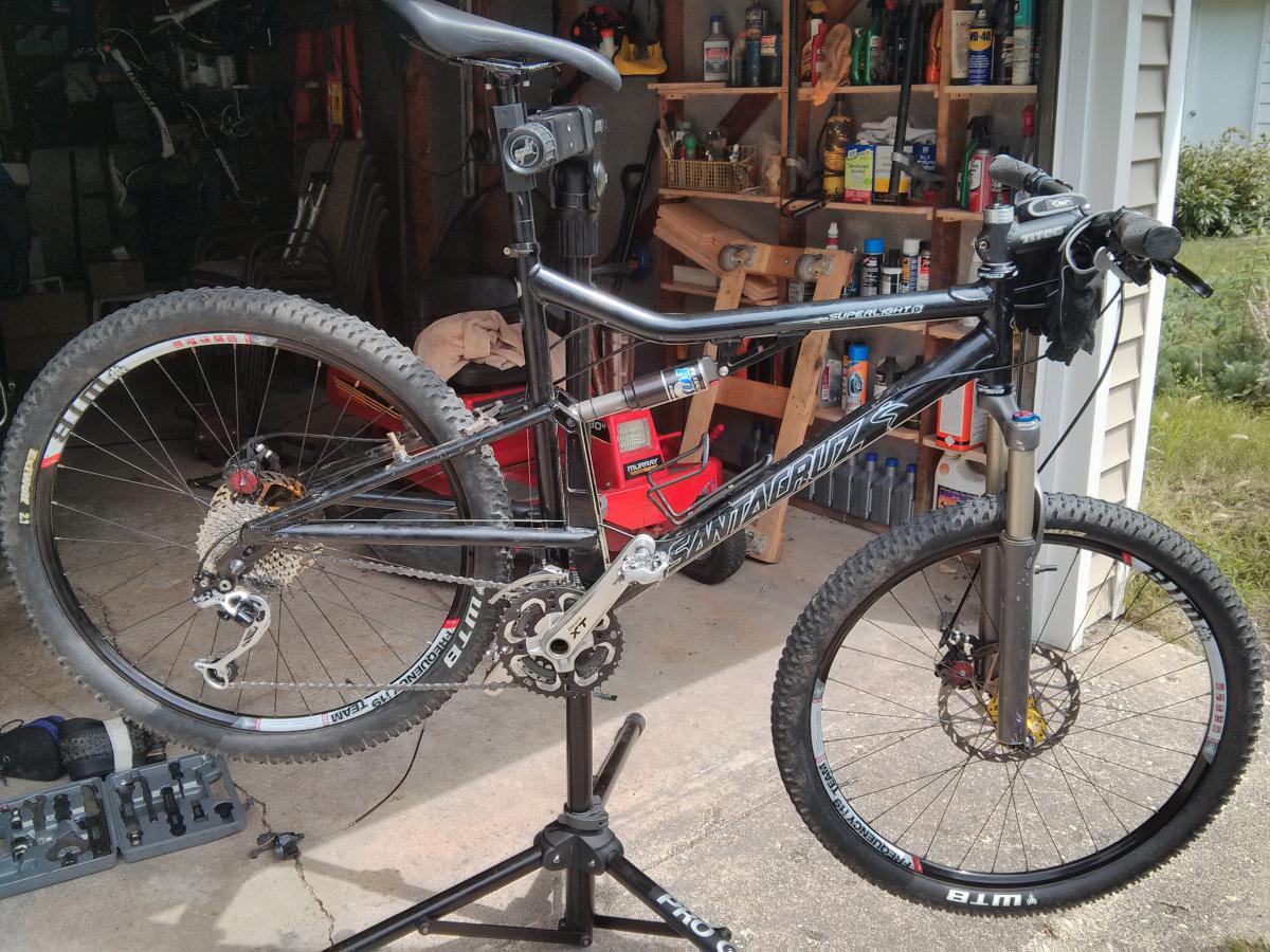 Santa Cruz Superlight: A black mountain bike mounted on a repair stand in a garage. The bike features a prominent suspension fork, a detailed drivetrain with a rear derailleur, and knobby tires. In the background, there are various tools and items stored on shelves, indicating a workshop environment.