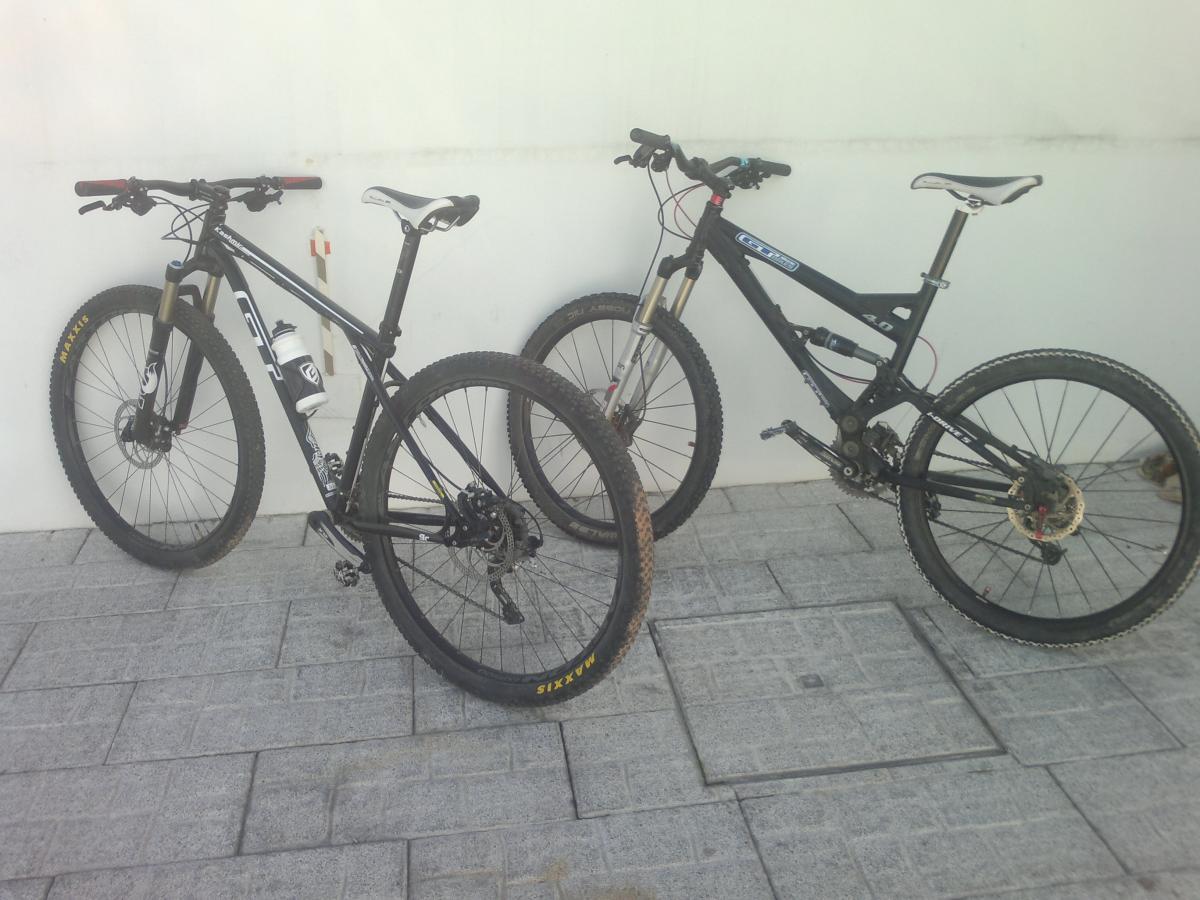 GT kashmir: Two mountain bikes are leaning against a white wall. The bike on the left is black with white accents and has a water bottle attached to the frame. The bike on the right is also black with a more angular design and visible suspension components. Both bikes have thick, knobby tires suitable for off-road riding and are positioned on a paved surface.