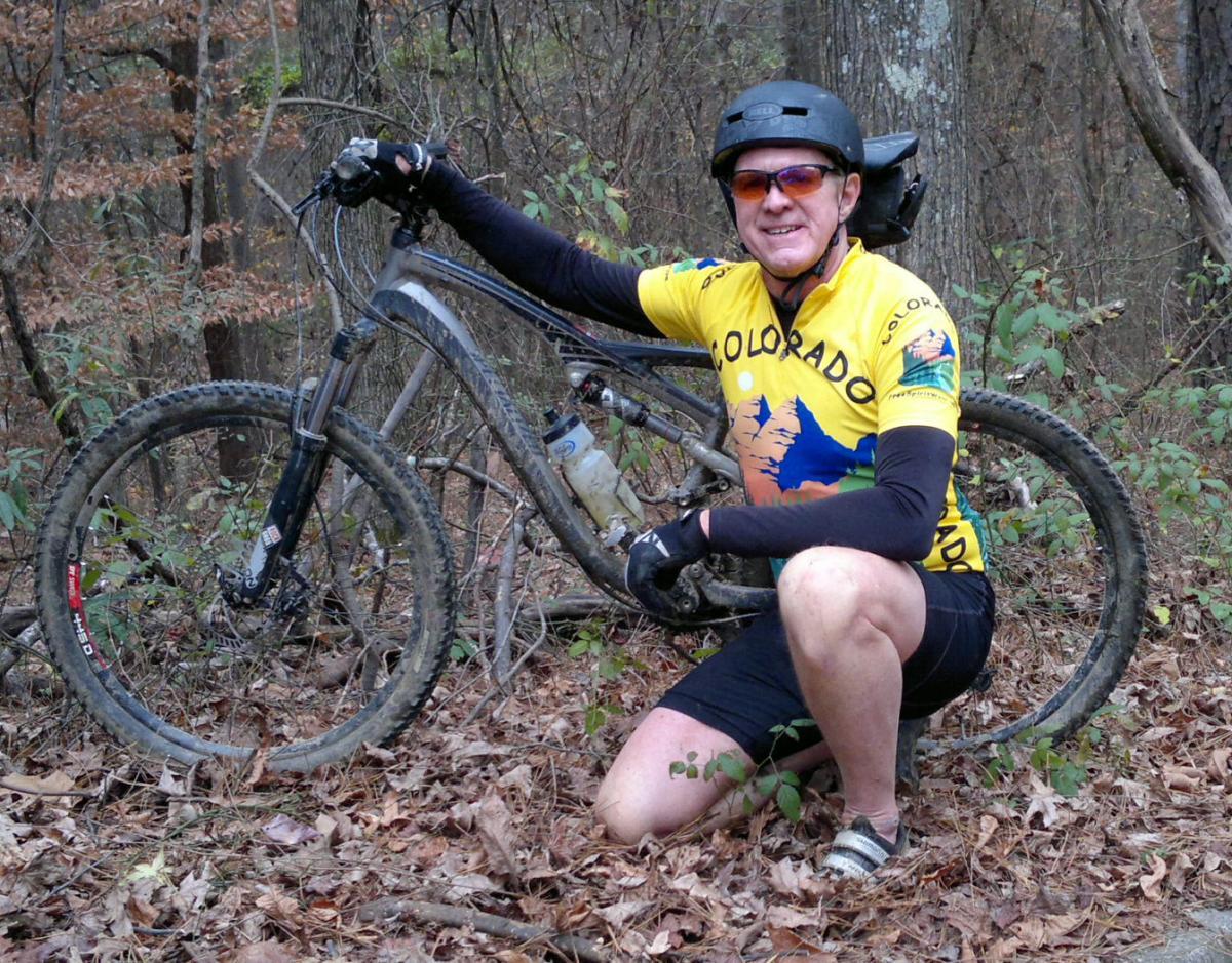 Specialized Camber: A person in a yellow Colorado-themed cycling jersey squats beside a mountain bike in a wooded area, surrounded by autumn leaves and trees. The cyclist, wearing a helmet and sunglasses, is smiling and appears to be enjoying the outdoor environment.