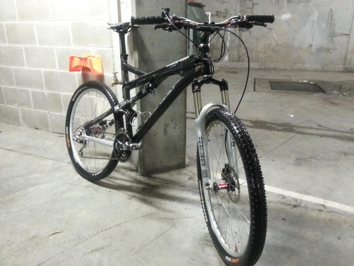 Titus Racer X Carbon: A mountain bike leaning against a concrete wall in a parking garage. The bike has a black frame with notable components, including a white front fork and knobby tires. In the background, there is a small orange object, and the ground appears slightly wet.