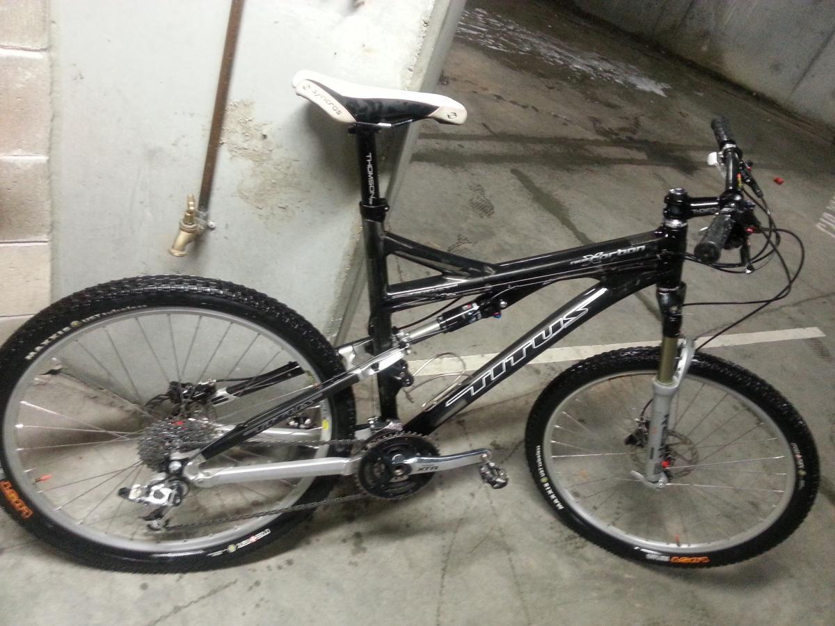 Titus Racer X Carbon: A black and silver mountain bike leaning against a wall in a parking garage. The bike features a dual suspension design, disc brakes, and knobby tires suitable for off-road riding. The floor nearby is concrete, showing signs of water, and there is a wall-mounted faucet in the background.