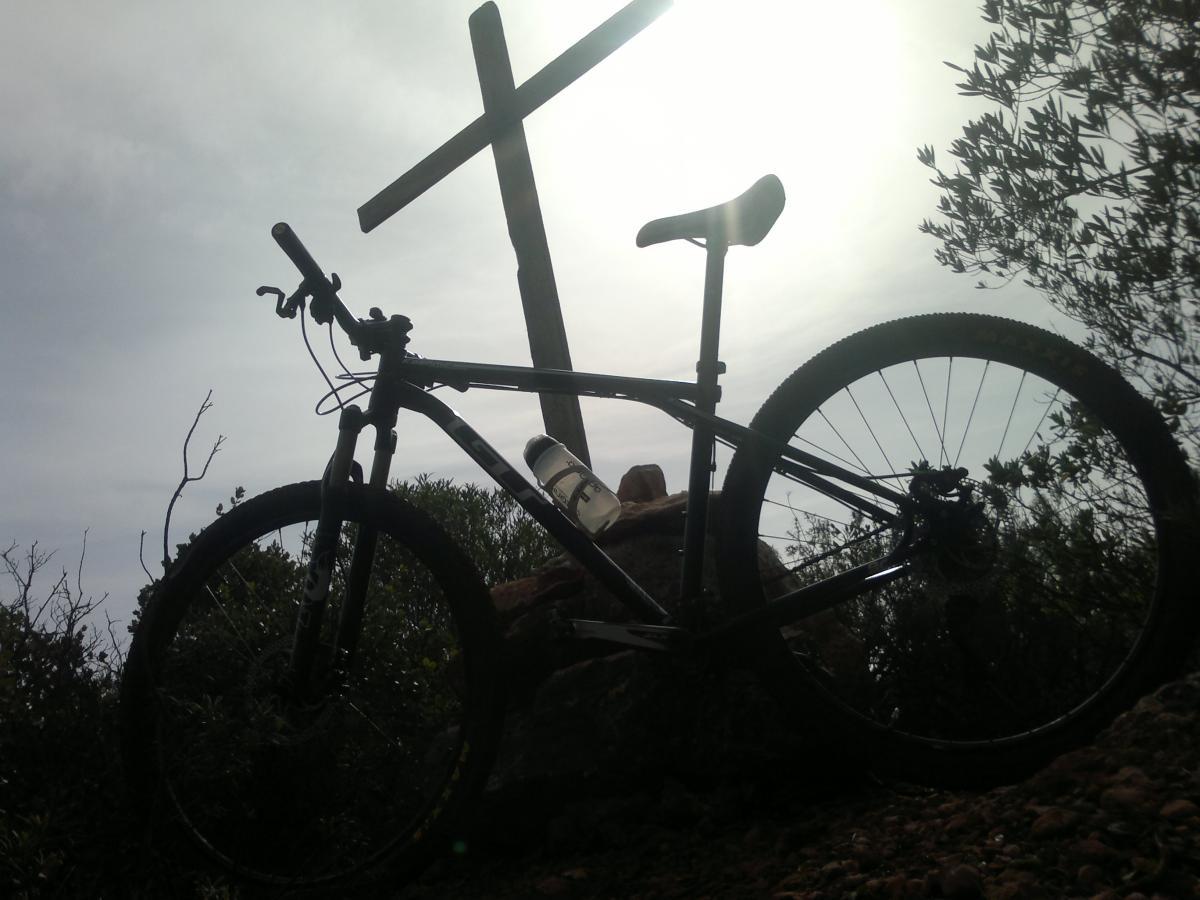 GT kashmir: Silhouette of a mountain bike resting on the ground with a wooden cross in the background. The image is taken outdoors, with a cloudy sky and some greenery surrounding the bike and cross.