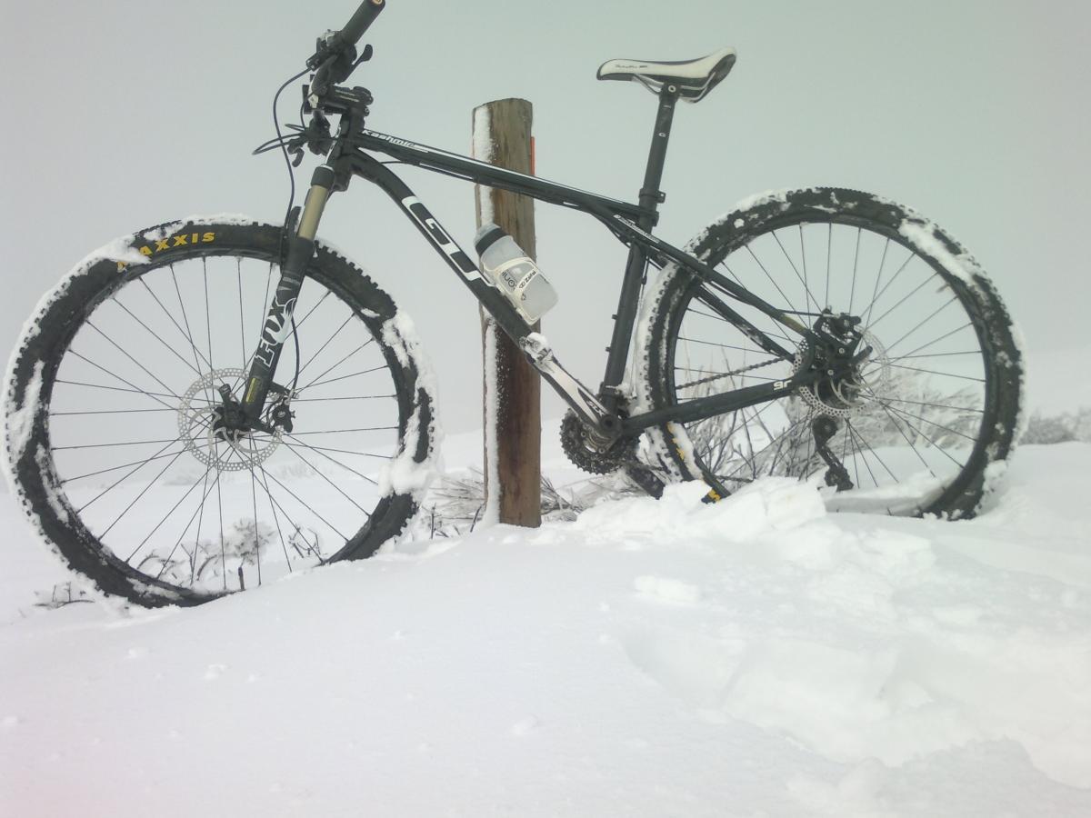 GT kashmir: A mountain bike resting on a snowy landscape, with snow covering the ground and accumulating on the tires and frame. A wooden post is positioned next to the bike, and the background is hazy due to fog or heavy snowfall. The scene emphasizes a winter outdoor environment.