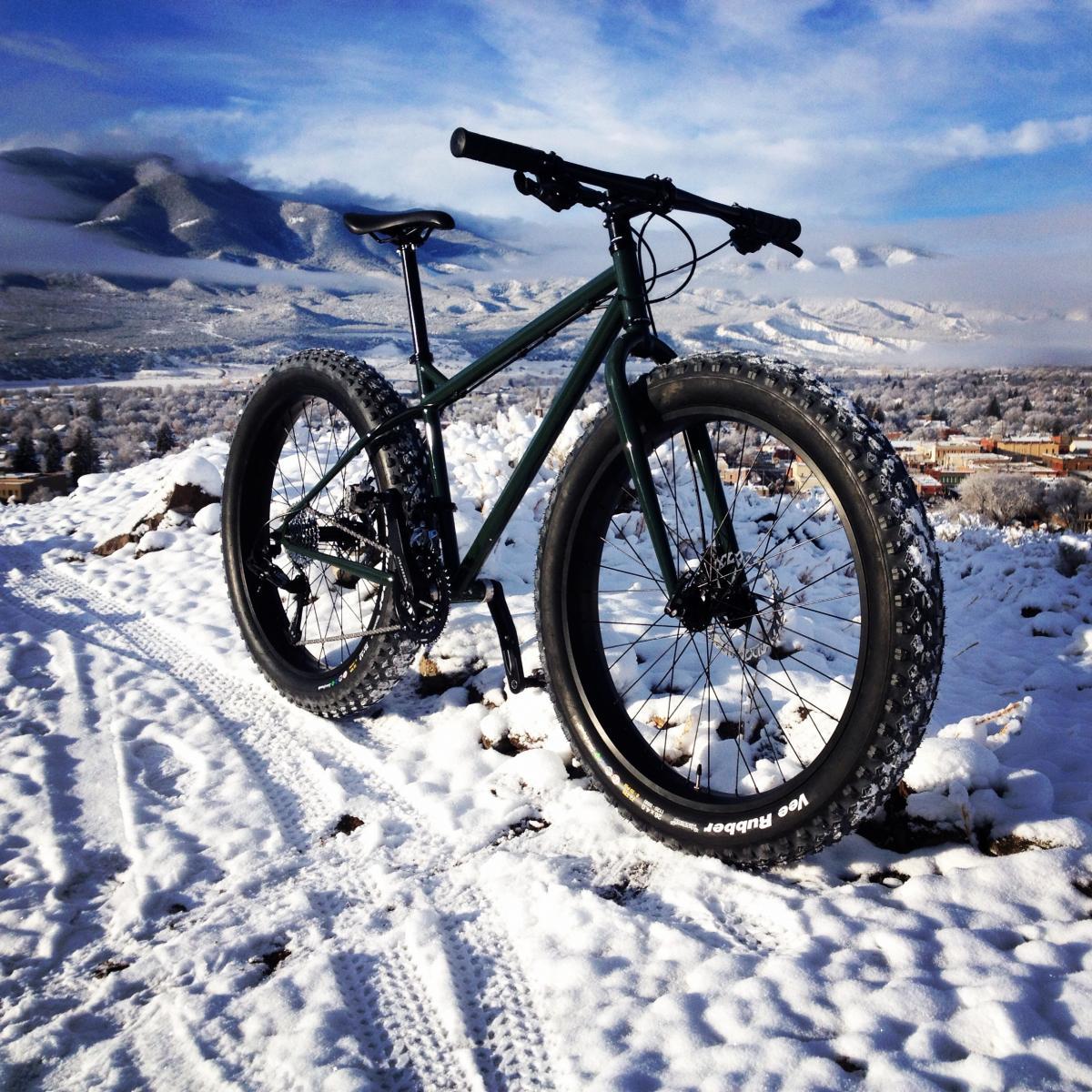A fat bike parked on a snowy trail with mountains in the background under a clear blue sky. The scene includes fresh snow covering the ground, with tire tracks visible in the snow. Arkansas Hills mountain bike trail.