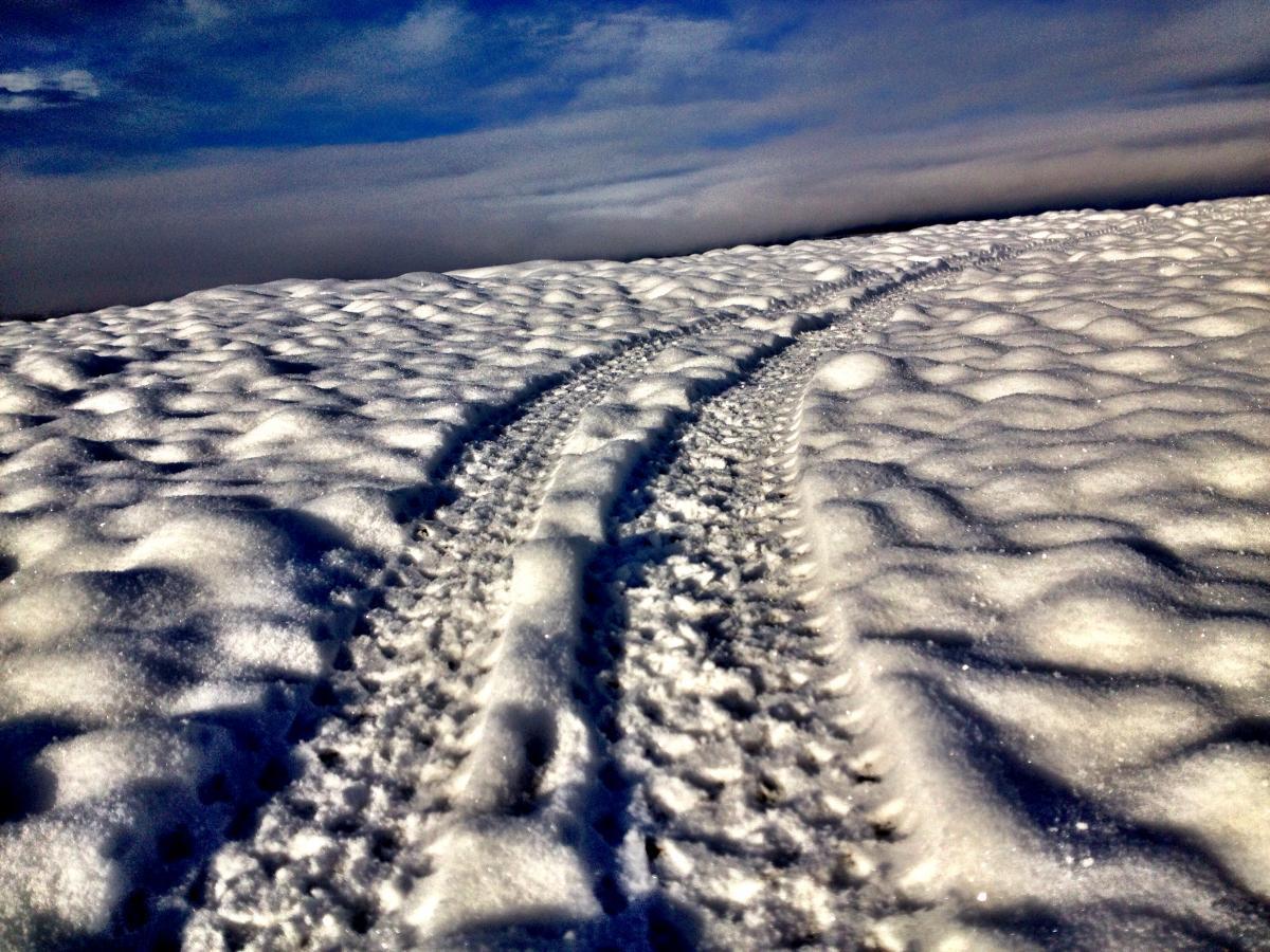 A snowy landscape with tire tracks winding through the thick blanket of snow, under a partly cloudy blue sky. The surface of the snow shows ripples and textures indicating movement, creating a serene winter scene. Arkansas Hills mountain bike trail.